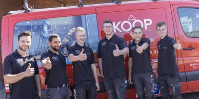 Sechs junge Männer in schwarzen T-Shirts stehen vor einem roten Lieferwagen und zeigen Daumen hoch, lächelnd und freundlich.