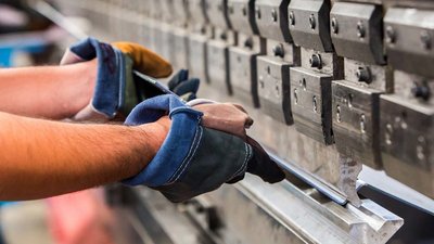 Detailaufnahme von Händen bei der Metallverarbeitung Ein Arbeiter mit Handschuhen hält ein Metallstück, während er eine präzise Bearbeitung an einer Maschine vornimmt.