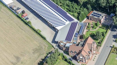 Eine Luftaufnahme zeigt ein modernes Gebäude mit Solarpanelen, umgeben von gepflegten Gärten und einem Feld.