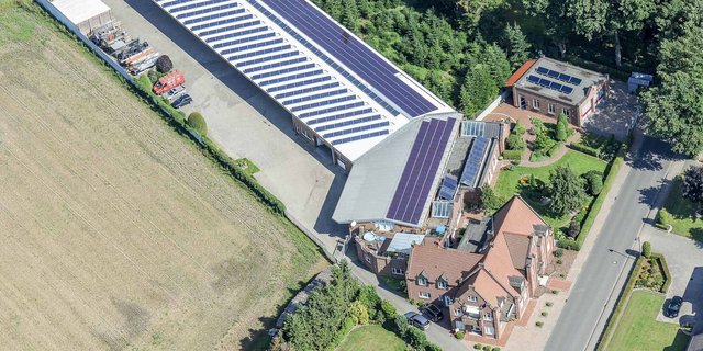 Eine Luftaufnahme zeigt ein modernes Gebäude mit Solarpanelen, umgeben von gepflegten Gärten und einem Feld.